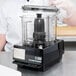 A person in a white coat and plastic gloves using a Waring S Blade in a black food processor on a white counter.