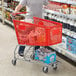 A man pushing a Regency red plastic grocery cart full of products down a grocery store aisle.