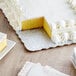 A yellow frosted cake with a slice missing on a silver Enjay corrugated cake board.