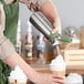 A person using a green stainless steel iSi whipped cream dispenser to pour whipped cream on a drink.