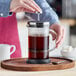 A person using an Acopa glass French coffee press to make coffee on a table in a coffee shop.