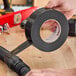 A roll of black Nashua duct tape being applied to a pipe fitting on a wooden workbench.