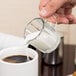 A person using an Anchor Hocking glass creamer to pour milk into a cup of coffee.