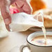 A hand pouring milk from an Acopa glass carafe into a cup of coffee.