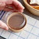 A hand holding a container of brown liquid with a Choice round PET take-out lid filled with chocolate pudding.