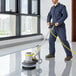 A man in blue overalls using a Lavex Pro single speed rotary floor machine to clean a floor in a professional kitchen.