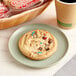 An individually wrapped Best Maid Chocolate Chip Cookie with M&M's on a plate next to a cup of coffee.