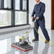 A man in a blue coverall using a Lavex Pro rectangular floor machine to clean a floor in a professional kitchen.