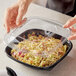 A hand placing a clear plastic dome lid on a Visions square bowl of food.