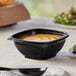 A black Visions plastic square bowl filled with soup on a table.