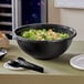 A bowl of salad in a black Visions plastic bowl with a black spoon on a table.