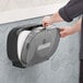 A person putting a double roll of Tork toilet paper into a black plastic dispenser.