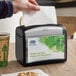 A person using a black Tork tabletop napkin dispenser on a counter.