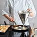 A person in a white coat using a de Buyer stainless steel piston funnel to pour pancake batter into a pan.