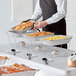 A man in a black apron serving food from a Choice chafer dish on a table with white and black striped tablecloths.