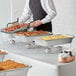 A man in an apron standing next to a hotel buffet with Choice disposable chafing dishes on a table.