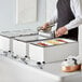 A man in an apron standing next to a hotel buffet with trays of food on table using Choice chafing dishes.