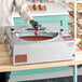 A person in a white coat and black gloves using a ChocoVision Revolation 3Z chocolate tempering machine to pour chocolate over a dessert.