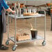 A stainless steel two-shelf utility cart with bottles, glasses, and a champagne bucket, being pushed in a restaurant setting.