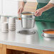 A person holding a silver pot of soup over a white Avantco drop-in well on a counter.