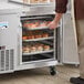 An unassembled aluminum sheet pan rack filled with food in front of a refrigerator.