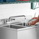 A person using a Regency deck-mounted faucet to wash dishes in a stainless steel sink.