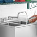 A person using a Regency deck-mounted faucet to wash dishes in a stainless steel sink.