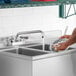 A person washing dishes in a stainless steel sink using a Regency deck-mounted faucet.