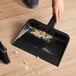 A black plastic utility dust pan being used to collect crumbs from a wooden floor.