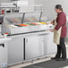 An Avantco countertop refrigerated prep rail on a white surface with food in containers. A man in a red apron and gloves is using it.