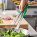 A hand holding a pair of Vollrath stainless steel tongs with green Kool-Touch handles over a bowl of broccoli.