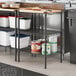 Regency black metal 3-shelf kit in a professional kitchen with wooden shelf inserts holding food containers.