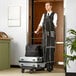 A man pushing a Lancaster Table & Seating black luggage cart with a black bag on top.