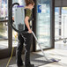 A woman using a ProTeam backpack vacuum with a white rectangular attachment to vacuum a carpet in a corporate office.