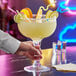 A hand holding an Acopa Colossal margarita glass of yellow liquid with fruit.