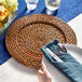 A hand holding a fork and knife on an Acopa rattan charger plate with a napkin