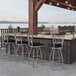 A group of Holland Bar Stool stainless steel outdoor extra tall bar stools with black seats next to a counter with a grill.