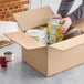 A person packing a Lavex heavy-duty cardboard shipping box with cans of food.