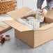 A person packing a Lavex Kraft cardboard shipping box with canned food.