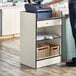 A person standing next to a Lancaster Table & Seating white cash register stand with a locking drawer.