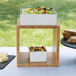 A white square table with a bamboo cube riser holding a container of pasta and salad.