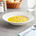 An Acopa wide rim ivory stoneware soup bowl on a table with a spoon and bread.