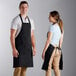 A man and woman standing on either side of a counter, both wearing black Choice standard bib aprons.