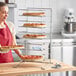 A woman wearing a red apron holding a tray of pizzas on a Choice oversized pizza pan rack.