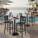 A black steel mesh outdoor bar stool with a ladderback design and a black wrinkle finish, shown by a poolside bar area.