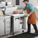 An apron-wearing man using gloves to put food into an Avantco 2 door sandwich prep refrigerator.