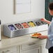 A woman standing at a counter with a ServSense stainless steel hotel pan organizer filled with condiments.