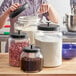 A woman using a scoop to fill an Acopa Dusk glass jar with brown granules.