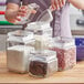 A woman pouring flour from a measuring cup into an Acopa clear glass jar.