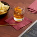 A bowl of chips and a glass of liquid on a table with a Choice Burgundy beverage napkin.